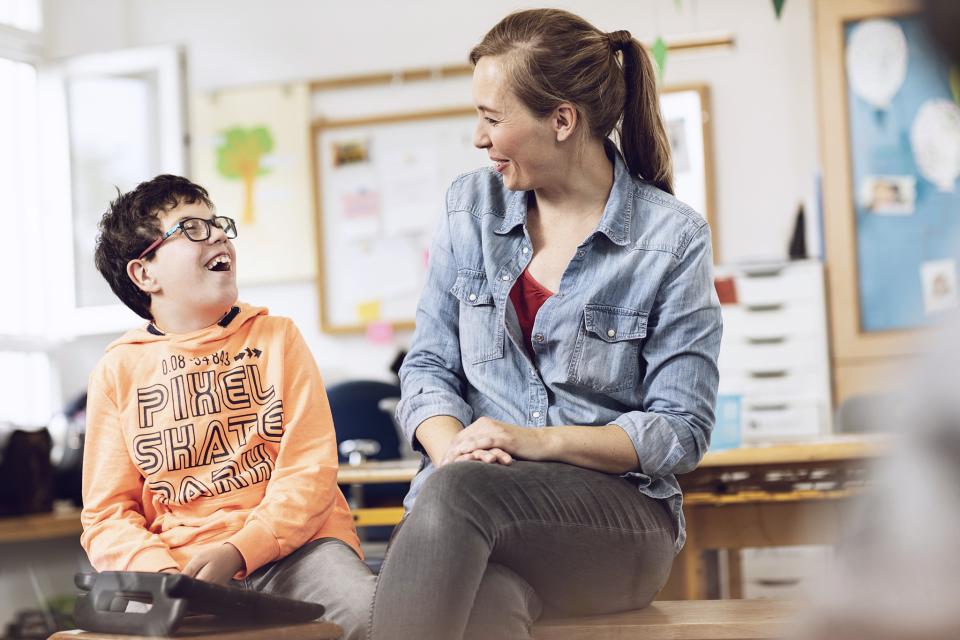 Eine Lehrerin sitzt und lacht mit einem Schüler mit sonderpädagogischem Förderbedarf.